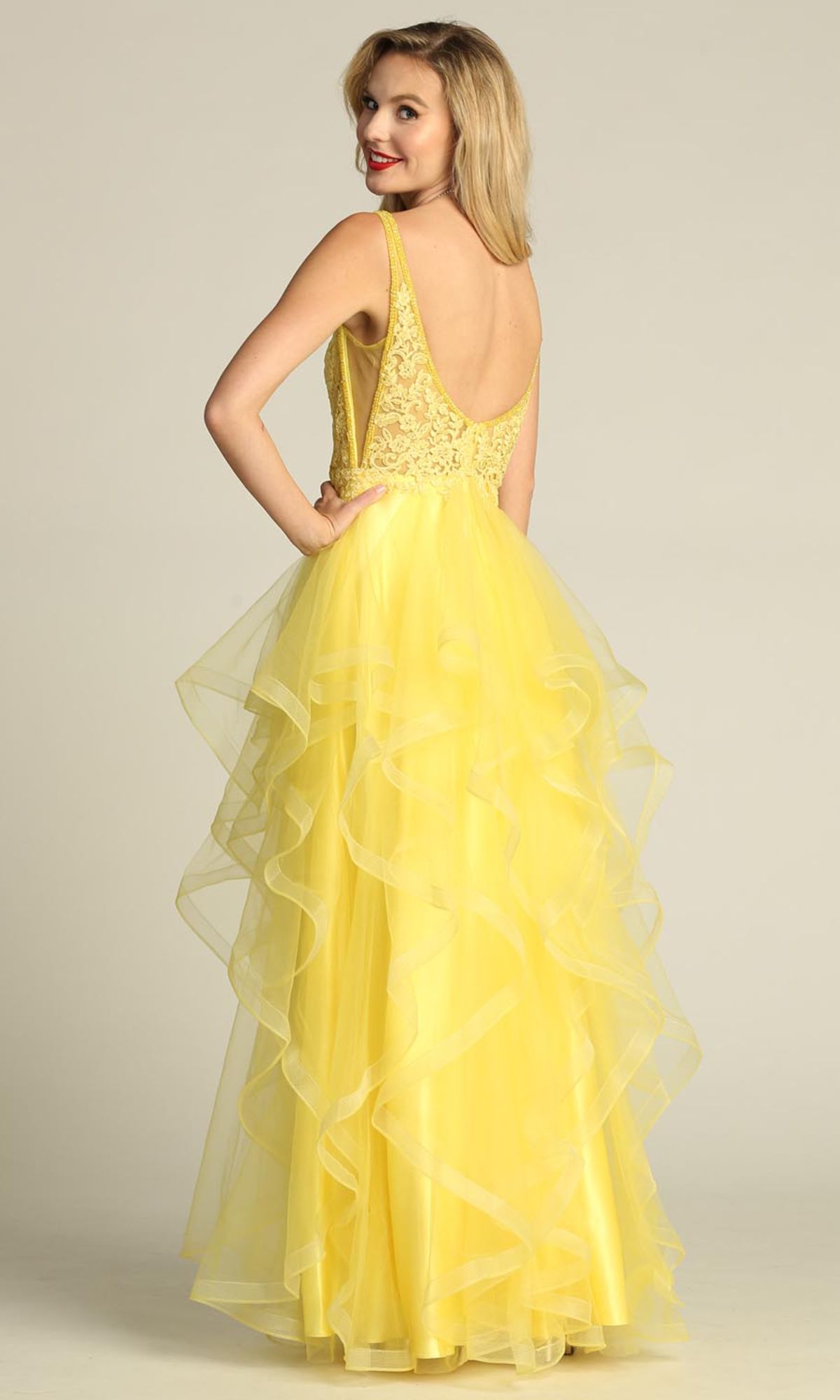 A woman wearing the Sheer-Sides Long Tiered Prom Dress (Chicas XR6006) with a lace bodice and layered tulle skirt stands facing away, looking over her shoulder and smiling.
